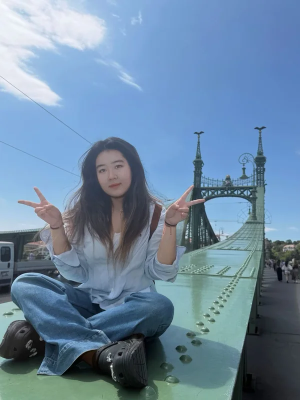 Anna sitting outdoors beside a bridge, making a peace sign.